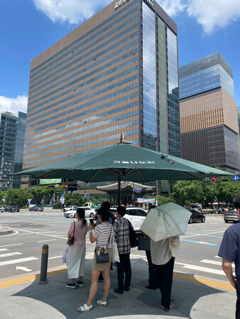 9日、ソウルの交差点に設置された大きな日傘の下で、信号待ちをする人々